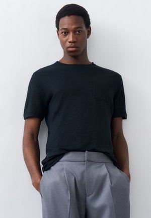 Νέος άνδρας με κοντά, σγουρά μαλλιά στέκεται μπροστά σε ανοιχτόχρωμο τοίχο, φορώντας μαύρο πουκάμισο με κοντά μανίκια και γκρι πλισέ παντελόνια, με τα χέρια στις τσέπες.