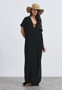 Fekete maxiruha V-nyakkivágással és bő ujjakkal, mindkét oldalon hasítékkal. Széles karimájú, bézs kalappal viselve.