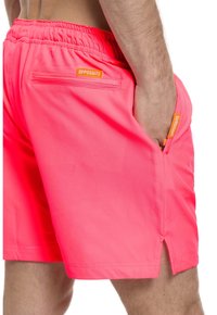 Rožiniai maudymosi šortai su elastine juostos juosta, šoninėmis kišenėmis ir užpakaline kišene. Juostos juostoje yra oranžinė prekės ženklo etiketė.