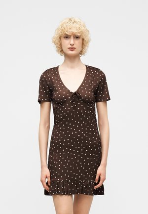 Personne blonde portant une robe marron à manches courtes avec des pois blancs et un ourlet à volants, debout face à l'avant sur un fond uni.