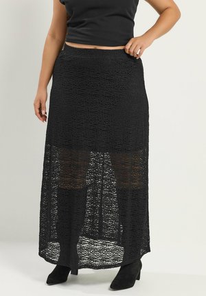 Mujer vestida con una falda maxi negra con textura y una sección inferior translúcida, y botas negras de tacón hasta el tobillo, posando frente a un fondo sencillo.