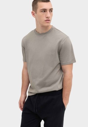 Young man wearing a light gray short-sleeve t-shirt and black pants, looking to his left with hands in pockets against a plain background.