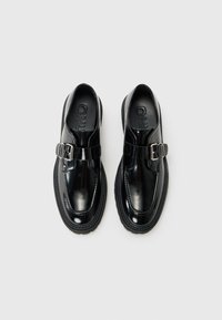 Zapatos negros pulidos que presentan una correa con hebilla, punta redonda, suela de goma plana y acabado brillante. Se aprecian detalles de costura sutiles.