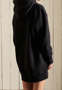 Mustapohjainen oversized-huppari, jossa on huppu, pitkät hihat, pehmeä tekstuuri ja ribbaremmitys. Takana on yksinkertainen pinta, jossa on vain minimikokoista ompelua.
