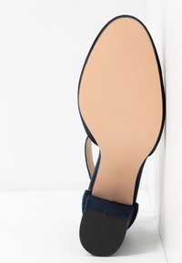 Semelle d'une chaussure de femme bleu marine à talon bas noir et semelle extérieure beige lisse sur fond blanc.
