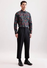 Langarm blouse met knopen van donkergroen stof met een bloemmotief. Gecombineerd met een hoog getailleerde, geplooide zwarte broek en schoenen.