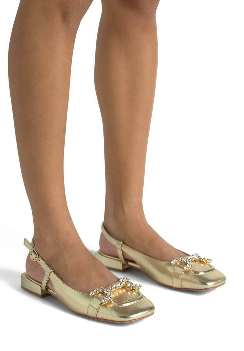 Ballerine metalliche dorate con punta quadrata e tacco basso. Accentuate da un motivo di cristalli sulla punta. Design con cinturino alla caviglia.