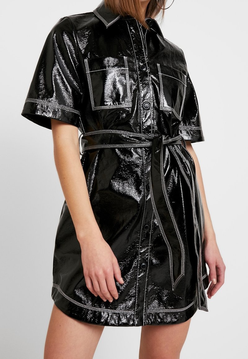 Svart, glänsande patenterad skjortklänning med korta ärmar, krage, två bröstfickor och cinchad midjebälte; har vita sömnadsdetaljer.