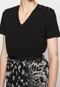 Schwarzes V-Ausschnitt-Oberteil mit kurzen Ärmeln und dekorativen Ösen an der Schulter. Die Bluse hat einen elastischen Bund und wird mit einem gemusterten Rock kombiniert.