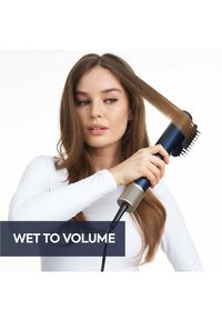 Frau benutzt eine blaue Haartrocknerbürste, um lange nasse braune Haare zu stylen, mit Fokus auf Volumen, trägt ein weißes langärmeliges Shirt.