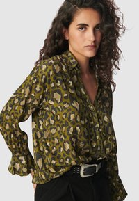 Oliwkowa bluzka z wzorem w panterkę w różnych odcieniach. Posiada zapięcie na guziki z przodu, długie rękawy i luźny krój.