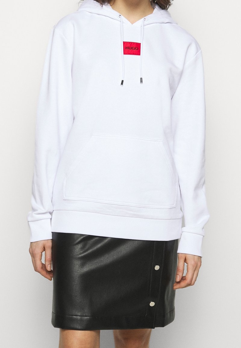 Sudadera blanca con bolsillo frontal y parche rojo del logo "HUGO", combinada con una falda negra de cuero sintético con acentos de botones a presión.