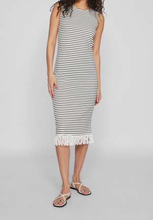Femme portant une robe midi blanche sans manches avec des rayures horizontales noires et un ourlet à franges, associée à des sandales blanches plates.