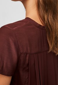 Blouse bordeaux à manches courtes, avec un design plissé à l'arrière et une texture lisse. Couture subtile le long du décolleté.