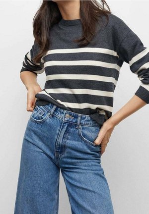 Personne portant un pull rayé gris foncé et blanc et un jean bleu taille haute, mains posées sur les hanches.