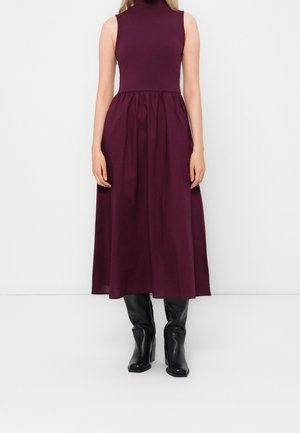 Sukienka midi bez rękawów z wysokim kołnierzem i plisami w ciemnym fiolecie, zestawiona z czarnymi, sięgającymi połowy łydki botkami na obcasie, stojąca na jednolitym białym tle.