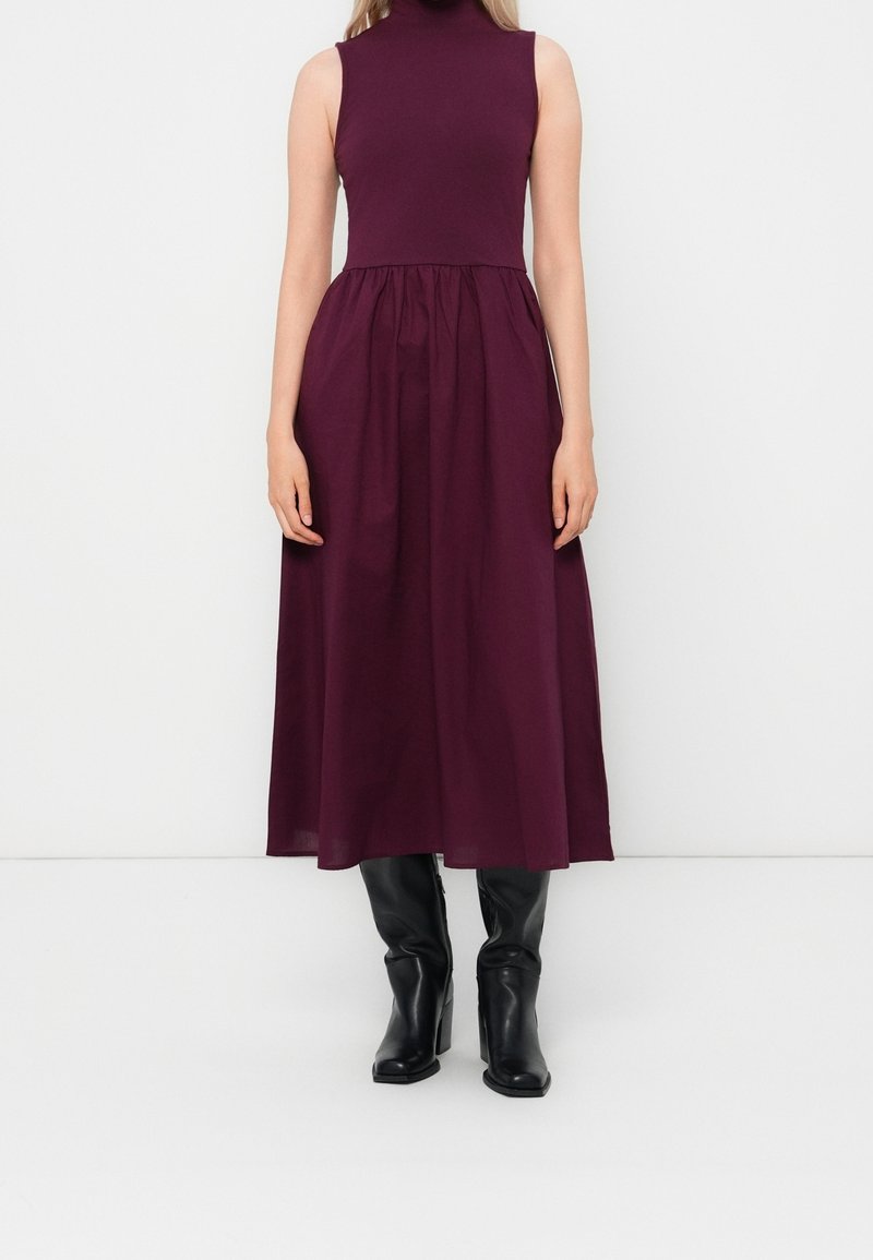 Robe midi plissée sans manches à col montant, en violet foncé, associée à des bottines noires à talons mi-mollet, posée sur un fond blanc uni.