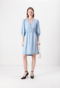 Rochie din denim albastru deschis, cu mâneci tip puff, decolteu în V și talie strânsă. Asortată cu pantofi cu toc înalt negri și o geantă roz.
