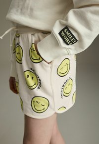 Krótkie, kremowe szorty z powtarzającym się wzorem żółtych uśmiechniętych buźka. Wzdłuż pasa zdobione czarną metką "SMILEY WORLD".
