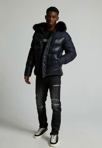 Černá péřová bunda s kapucí lemovanou kožešinou, se zipem a vodorovnými švy. Kombinována s opranými tmavými džínami a černými teniskami.