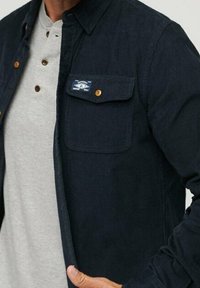 Marine corduroy overhemd met knoopsluiting, enkele borstzak met label, gedragen over een lichtgrijze henley met houten knopen.