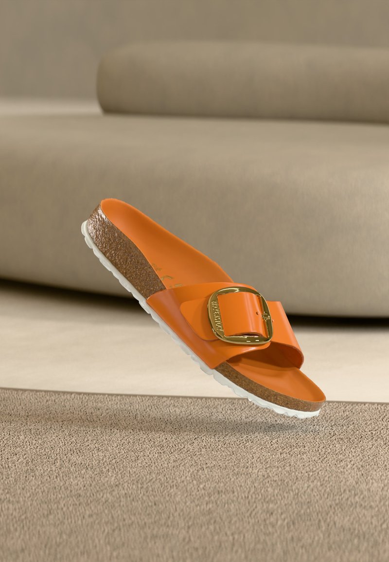 Πορτοκαλί σανδάλι τύπου slip-on με μεγάλη χρυσή αγκράφα, φελλό πάτο και λευκή ραβδωτή σόλα, τοποθετημένο σε φόντο μπεζ χαλιού και καναπέ.