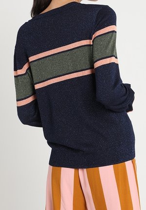 Kvinde iført en navyblå sweater med lyserøde og grønne vandrette striber og vidde bukser med lodrette lyserøde og sennepsstriber.