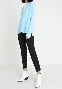 Lichtblauwe gebreide trui met V-hals, gedragen over een witte top, gecombineerd met zwarte slim-fit broeken en witte enkellaarsjes met een wiggenhak.