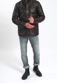 Mann trägt eine dunkelbraune Lederjacke, hellblaue hochgekrempelte Jeans und schwarze Stiefeletten. Er steht mit gefalteten Händen.