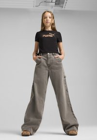 Camiseta negra con logo, combinada con jeans de pierna ancha en gris desvanecido. Presenta grandes bolsillos y un ajuste informal. Calzado marrón completa el look.