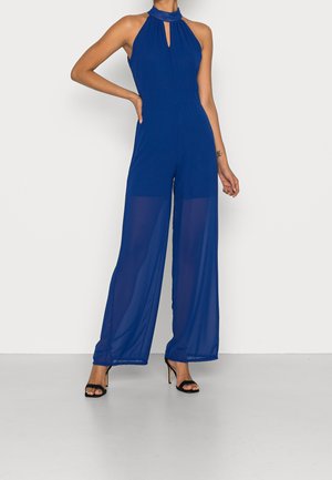 Koninklijk blauwe halter-neck jumpsuit met sleutelgatdetail, gemaakt van lichtgewicht stof. Kenmerkt zich door een wijdvallend ontwerp en doorschijnende panelen. Zwarte hoge hakken.