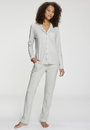 Woman standing barefoot wearing light grey button-up long-sleeve pajama top and matching pants with one hand in pocket.