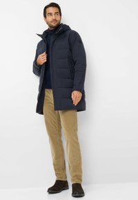 Cappotto imbottito blu navy con cappuccio, chiusura con zip e tasche laterali; indossato sopra un maglione scuro e abbinato a pantaloni in corduroy color beige e stivali scuri.