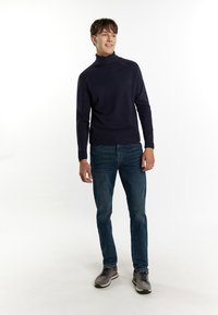 Maglione a collo alto di un blu navy scuro con polsini e orlo a coste, abbinato a jeans blu sbiaditi e scarpe da ginnastica grigie con accenti neri.
