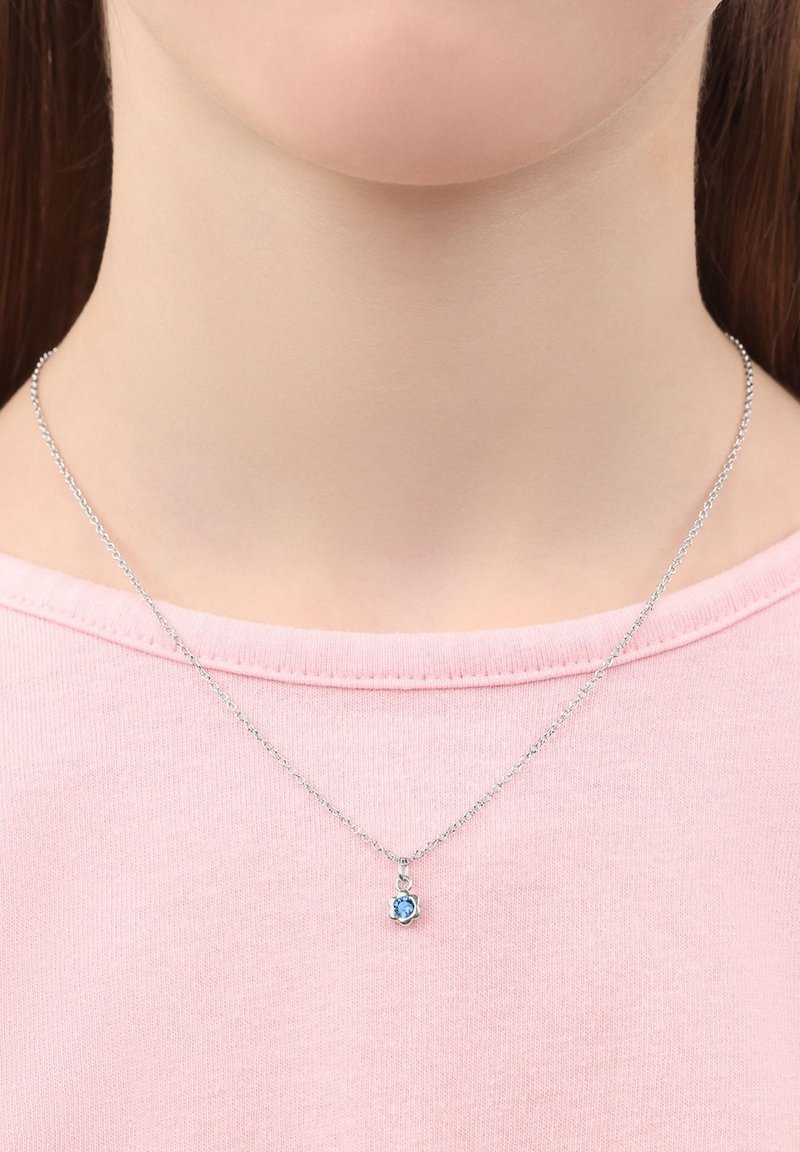 Silverkedja med ett hänge av blå ädelsten, bärs av en person i en rosa skjorta. Hänget har en enkel blommig design.