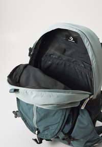 Grön och grå ryggsäck med rullöppning, texturerad tyg, dragkedjehuvudfack och intern varumärkesetikett. Öppen interiör synlig.