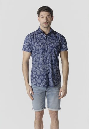 Hombre vestido con una camisa azul de manga corta con botones y estampado de hojas blancas, y pantalones cortos de mezclilla clara remangados, de pie frente a un fondo liso.