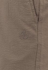 Tissu texturé marron avec logo brodé près d'une couture et de l'ouverture de la poche.