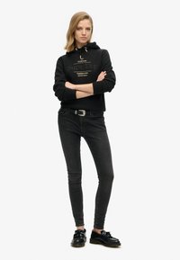 Sudadera negra con el logo "SuperDry", combinada con jeans skinny gris oscuro y zapatos negros. El conjunto presenta un diseño minimalista y una textura suave.
