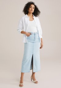 Fustă din denim albastru deschis cu o deschidere frontală, purtată cu un top alb fără mâneci și o cămașă albă desfăcută, completată cu pantofi cu toc deschis.