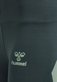 Ciemnozielone legginsy o gładkiej fakturze i dopasowanym kroju, z jasnym logo "hummel" umieszczonym po jednej stronie.