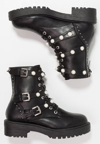 Stivali da combattimento in pelle nera con suole spesse, decorati con fibbie argentate, bottoni simili a perle e piccoli chiodi metallici, mostrati affiancati.