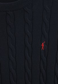 Marineblauwe kabelgebreide trui met verhoogde, gedraaide patronen en een klein rood logo van een golfer aan de linkerkant. Dikke ribgebreide halslijn.