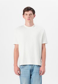 White short-sleeve T-shirt with a crew neckline, featuring a subtle text detail near the collar and a relaxed fit. Worn with light blue jeans.
