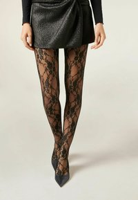Sorte blonderet strømpebukser med blomstermønster og små ædelsten, parret med en struktureret sort mini-nederdel og spidse sorte hæle.