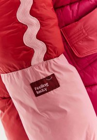 Rukáv červené a růžové bundy s vlnitým lemem a vyšitým štítkem, na kterém je nápis "Cítím se šťastně" a malá ilustrace zavřeného oka.