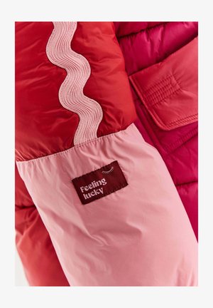 Rukáv červené a růžové bundy s vlnitým lemem a vyšitým štítkem, na kterém je nápis "Cítím se šťastně" a malá ilustrace zavřeného oka.