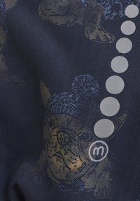 Marinfärgad tyg med fiskbensmönster som har abstrakta ansikten i guld och blått, med grå cirkulära detaljer och en liten logotyp längst ner.