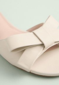 Sandale en cuir beige avec un large nœud sur le dessus. Design à bout ouvert et texture lisse ; semelle intérieure rose clair et petit talon minimaliste.