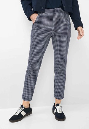 Persona in piedi che indossa pantaloni slim blu navy a motivo, sneaker nere e bianche e una giacca blu navy con una mano in tasca.